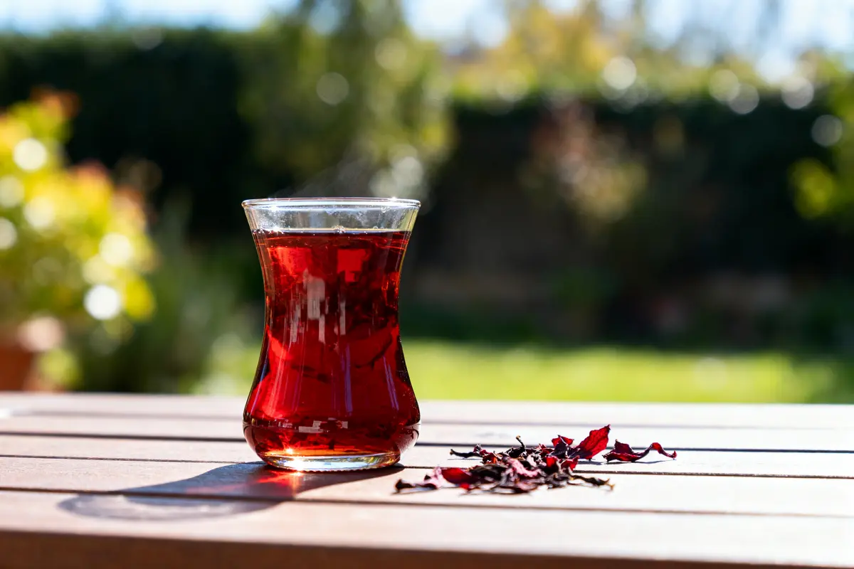 Verre de thé rouge sur table en extérieur, feuilles séchées à côté, fond flou verdoyant ensoleillé.
